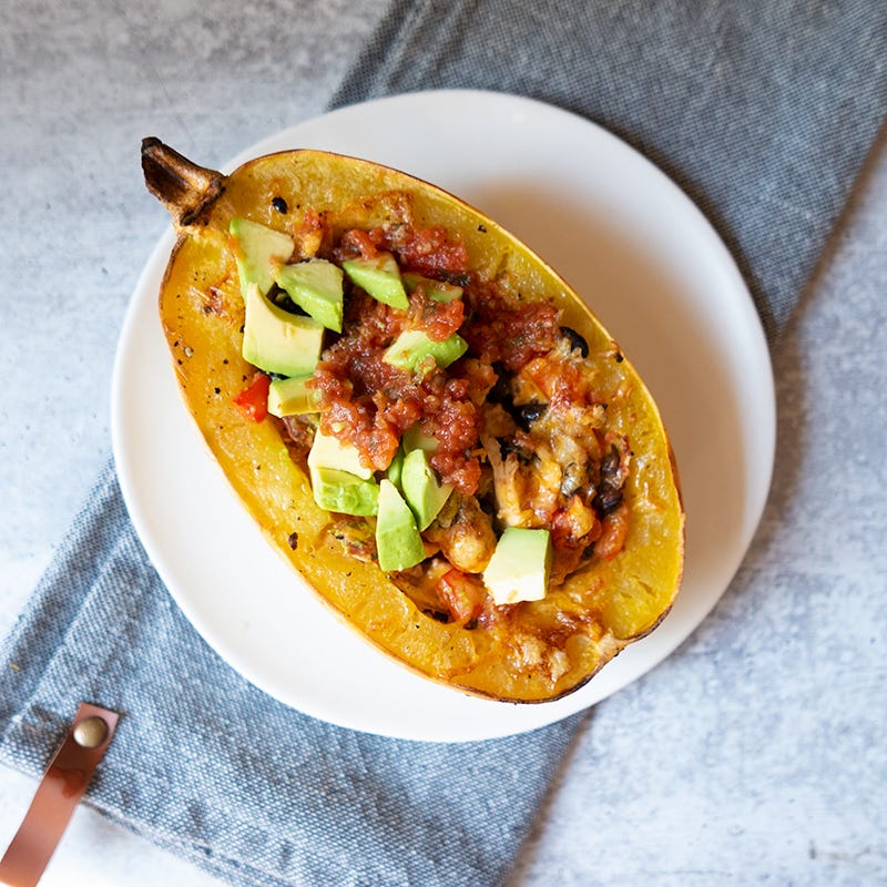 Roasted spaghetti squash half on a white plate filled with a cheesy chicken and bean mixture and topped with chopped avocado