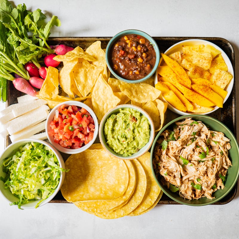 Large sheet pan topped with small bowls of chicken, lettuce, tomato, guacamole, salsa, and fresh fruit, plus some tortilla chips, fresh veggies, and tortillas