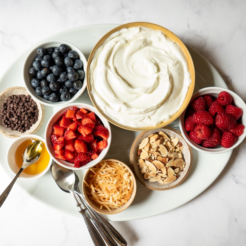 Large oval platter topped with small individual bowls of yogurt, fresh berries, nuts, coconut, chocolate chips, and honey