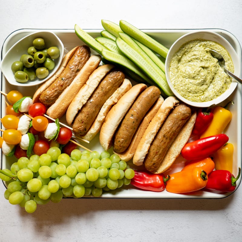 Large sheet pan topped with chicken sausages in buns, small bowls of olives and creamy pesto sauce, grapes, baby peppers, and tomato and mozzarella skewers