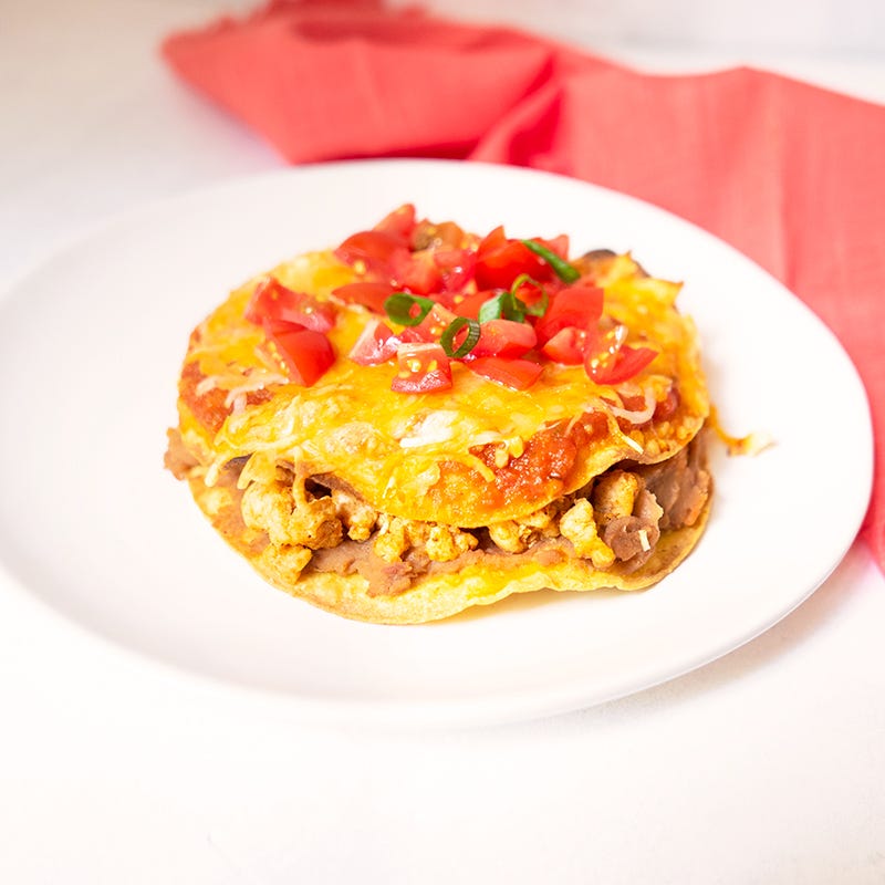 A small white plate topped with 2 toasted corn tortillas that are sandwiched with refried beans and seasoned turkey, then topped with pizza sauce, red salsa and melted cheese